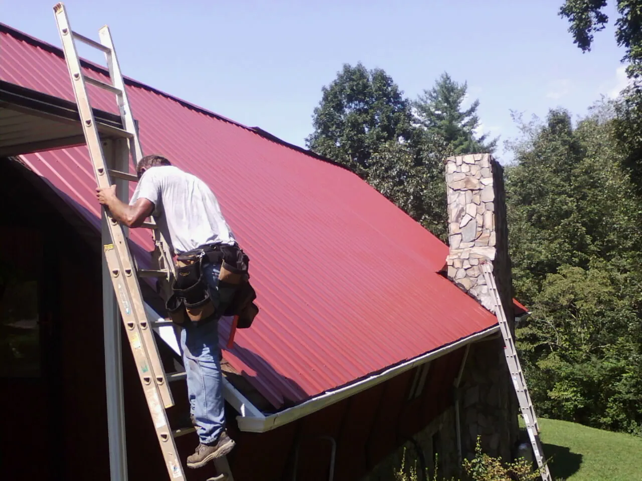 Roofer on a ladder beside a red metal roof and stone chimney — Soffit & Fascia Repair by Roofing Brothers in Broomall